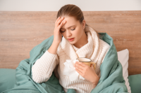 woman in bed with one hand on forehead and holding mug and a tissue in other hand