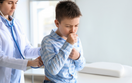 young boy sitting on exam table coughing while doctor checks his respiration