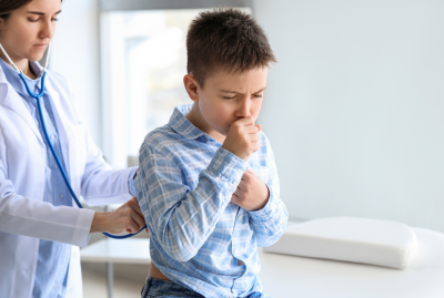 young boy sitting on exam table coughing while doctor checks his respiration